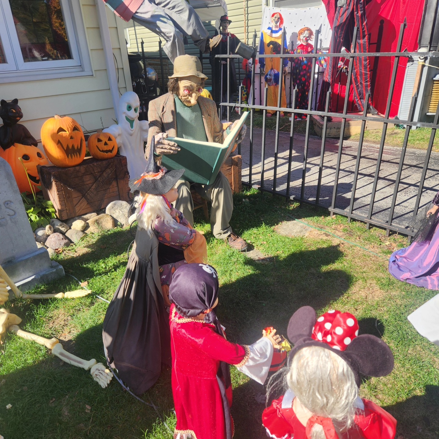 A Crypt Keeper figure reading to costumed child-sized figures behind cemetery fencing — Hills of Unrest, October 2024.