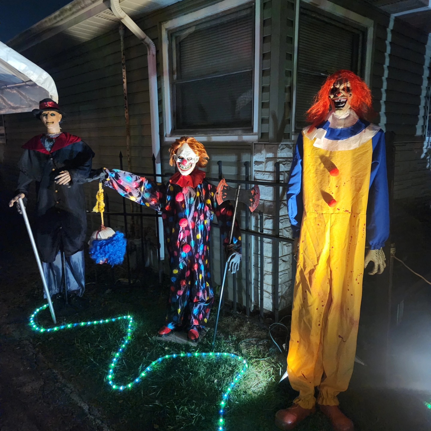 Three sinister clown figures beside the house, with glowing green rope lighting — Hills of Unrest, October 2024.